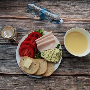 Zdjęcie przedstawia Dietę łatwostrawną: Kawa zbożowa z mlekiem kasza kukurydziana Bułka pszenna długa krojona Masło extra Szynka tostowa Zielona pasta jajeczna Pomidor Rukola Woda mineralna
